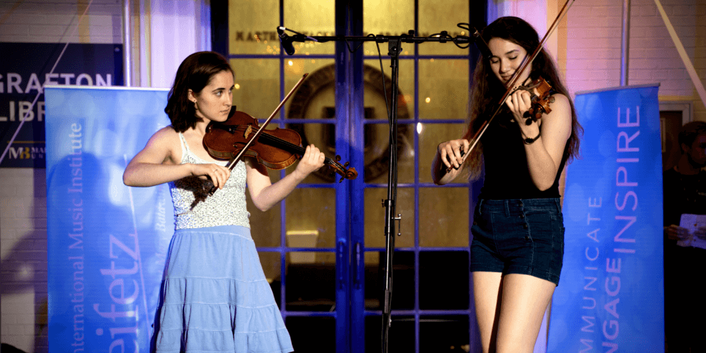 Two girls playing violin on a stage with spotlights shining on them