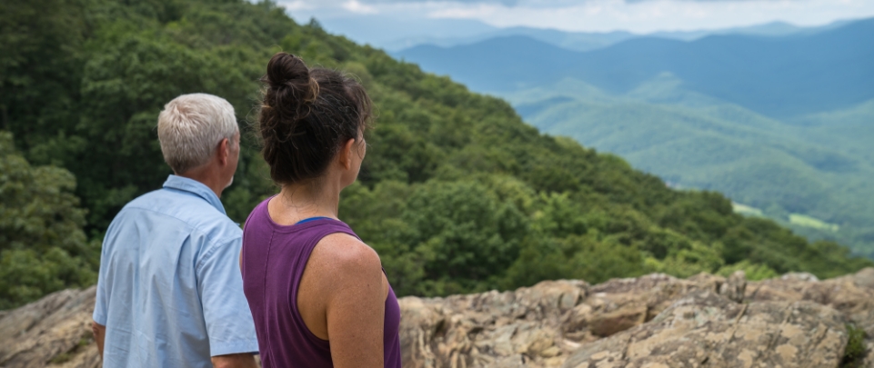 A Male And Female Couple Overlooks Mountains And Hills Covered In Greenery. 