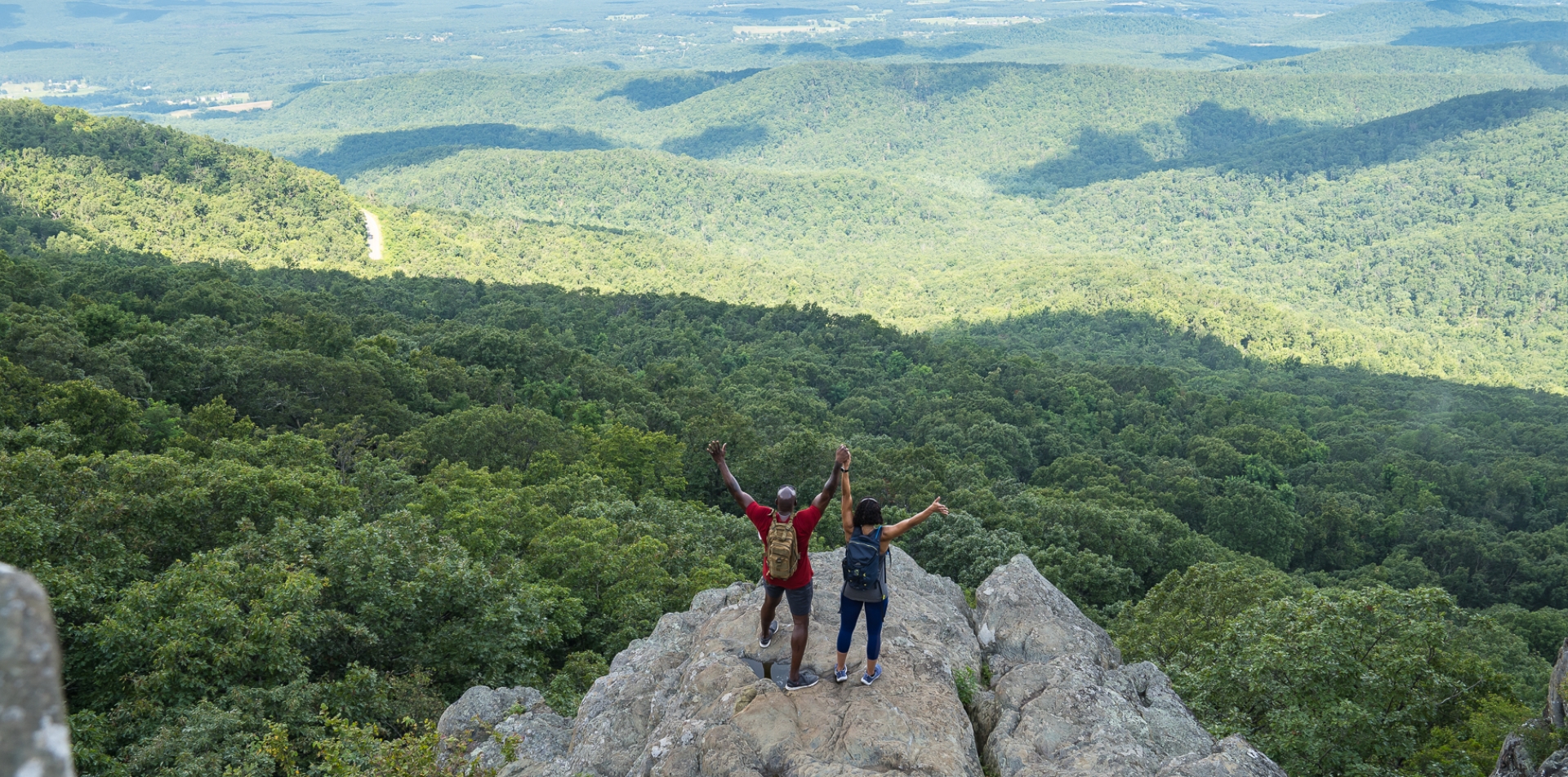 A couple holds hands with arms outstretched in the air while standing atop a mountain that overlooks hundreds of acres of greenery.  