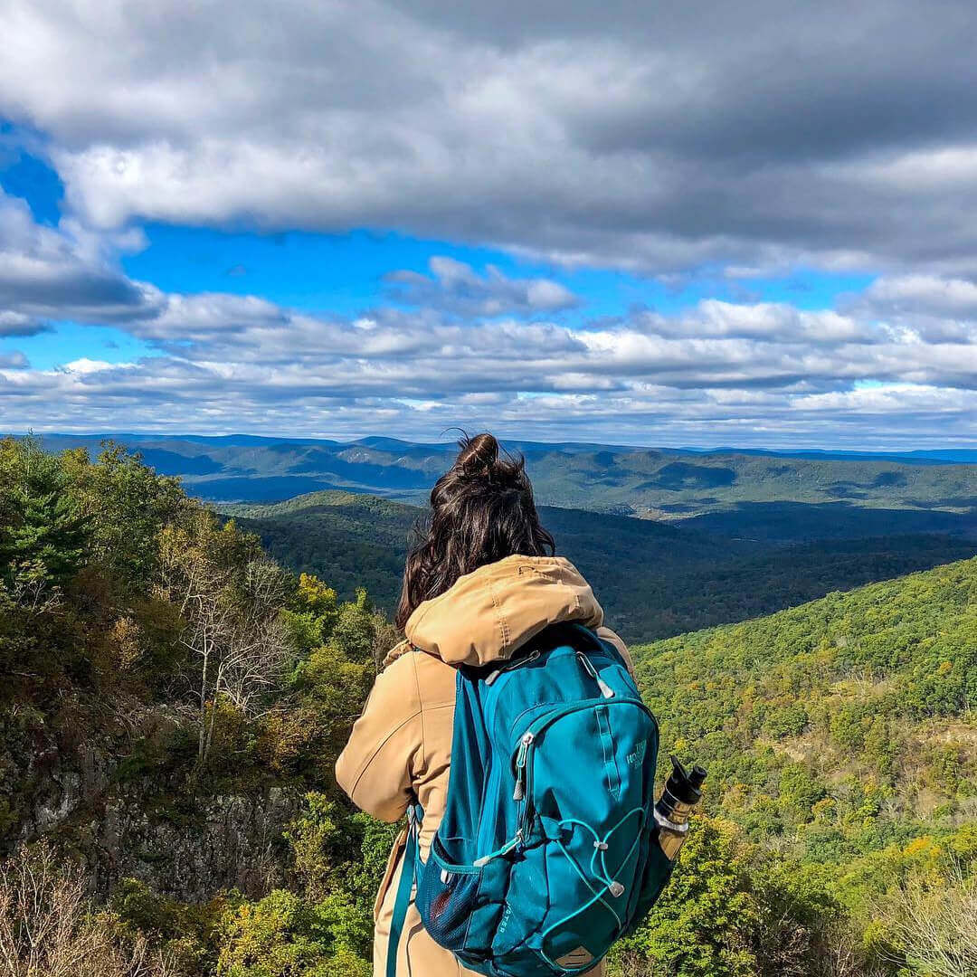 A back view of of a woman wearing a backpack looking out on to a view of mountains and hills. 