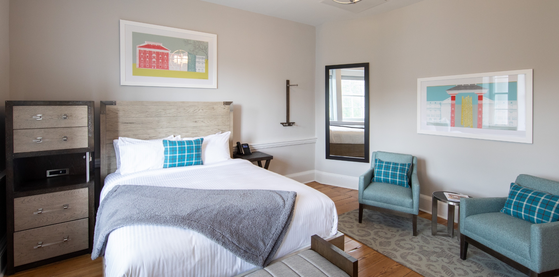 A guest room at the Blackburn Inn in Staunton Virginia is decorated with light wood furniture, blue chairs and white linens. Colorful paintings and a full size mirror hang on the wall. 