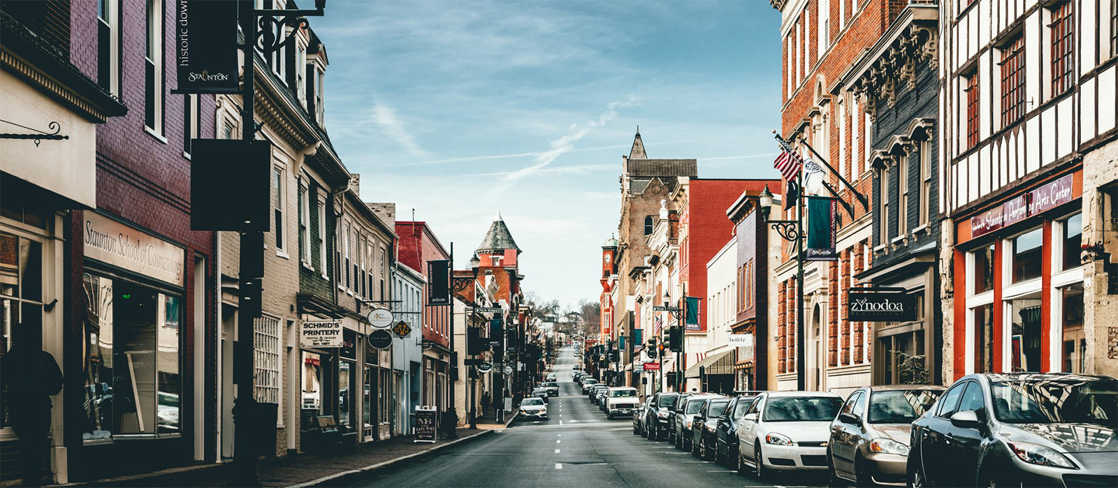 Street lined with cars, flags flapping in the breeze, and bustling energy in Downtown view of Staunton, VA 