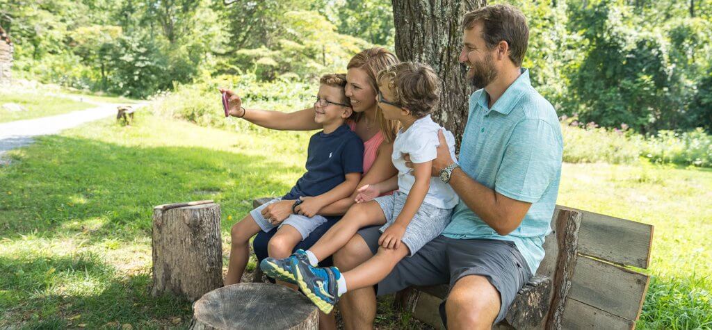 A family sitting on a bench taking a selfie under a tree on a summer day in staunton