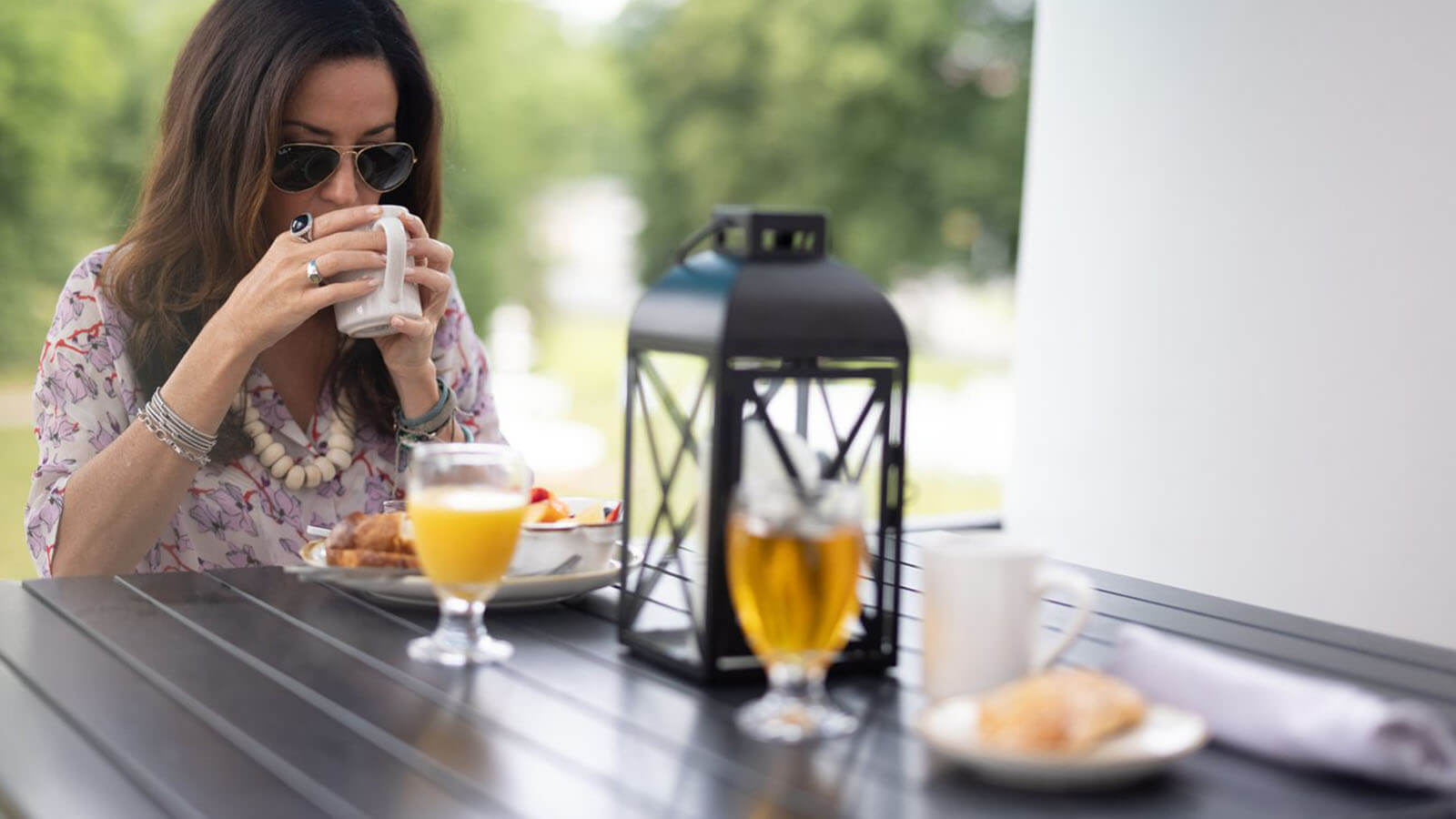 A woman sips coffee at an outside table with a breakfast platter in front of her. 