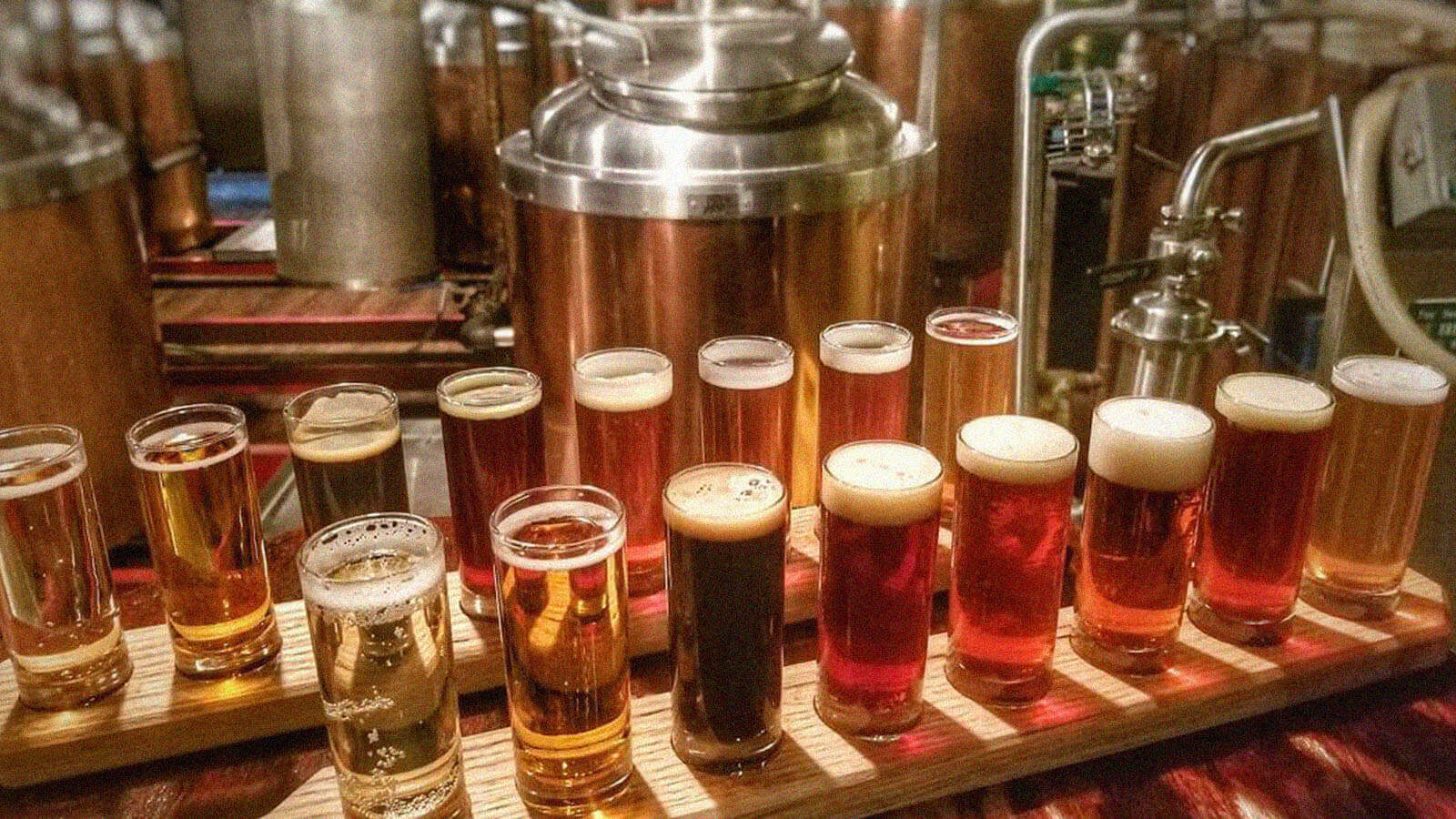 A flight of craft beers are lined atop wooden pillars in front of brewing equipment. 
