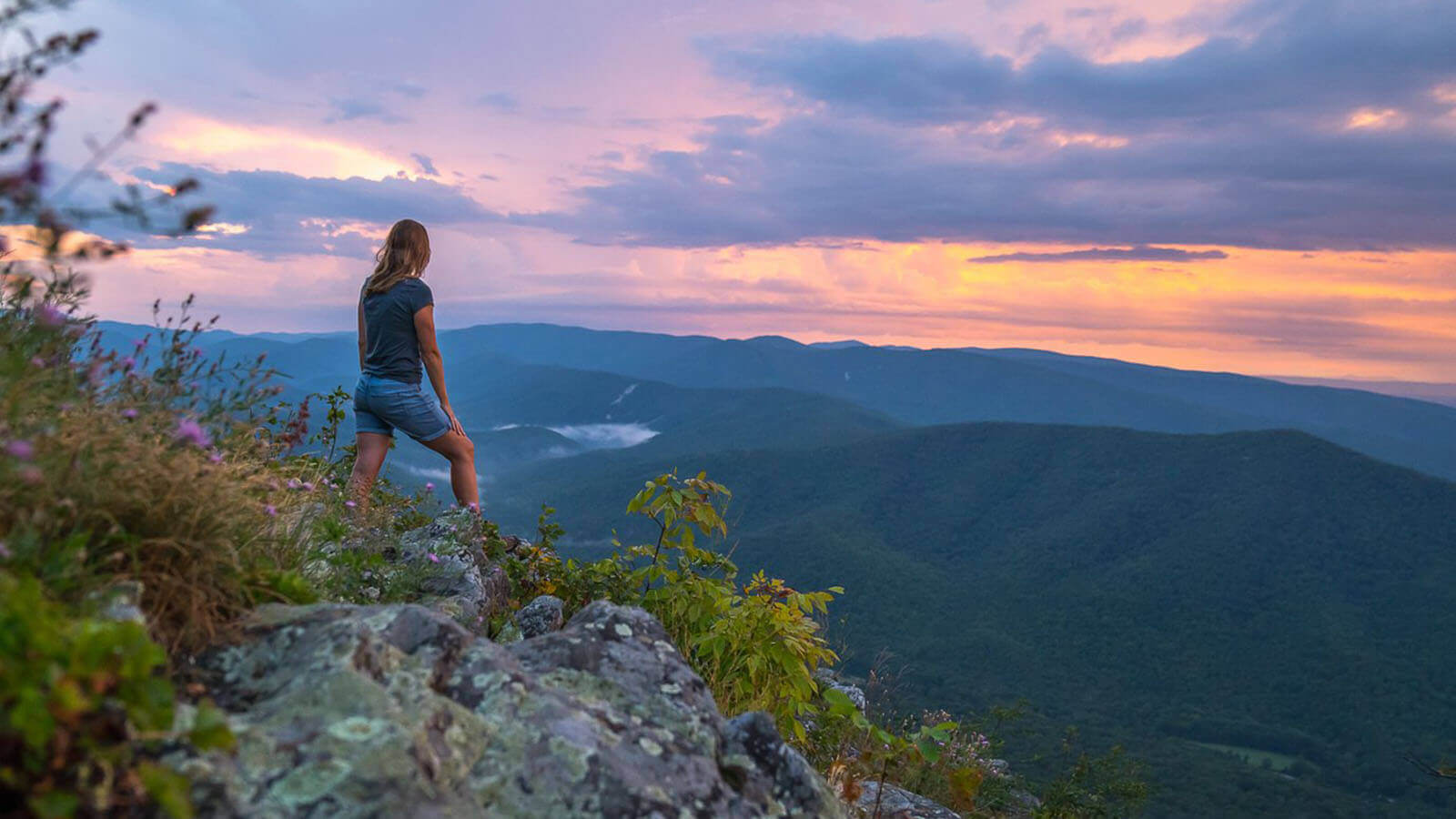 A female hiker stands atop a mountain looking at a blue and pink sunset over a valley of other mountains. 