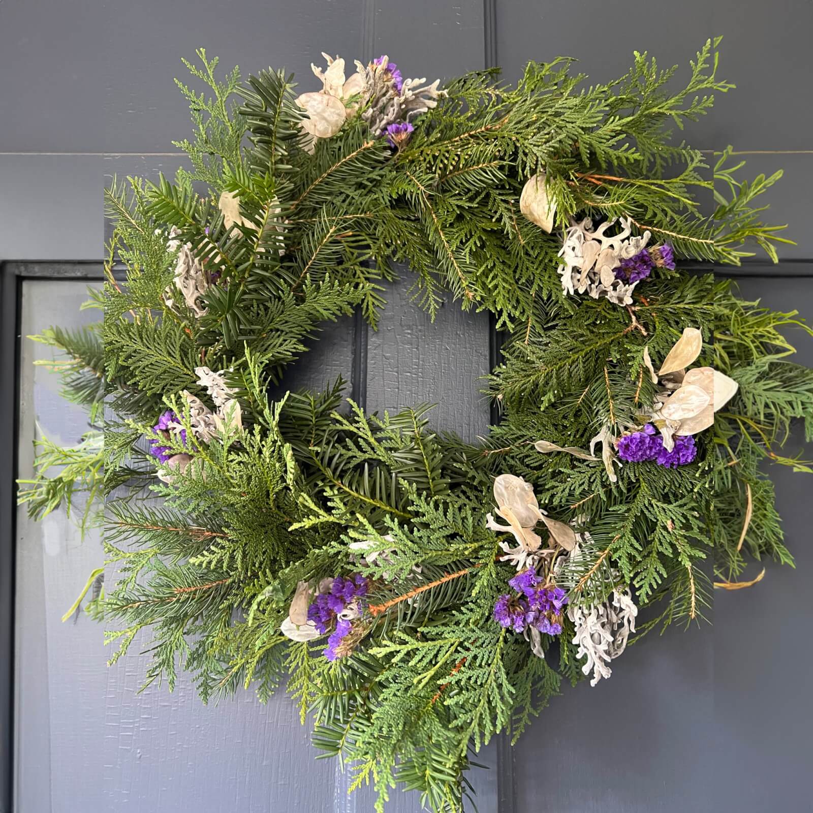 A Christmas wreath with purple and white dried flowers on a gray door made in Quince & Burdock's Wreath Making event