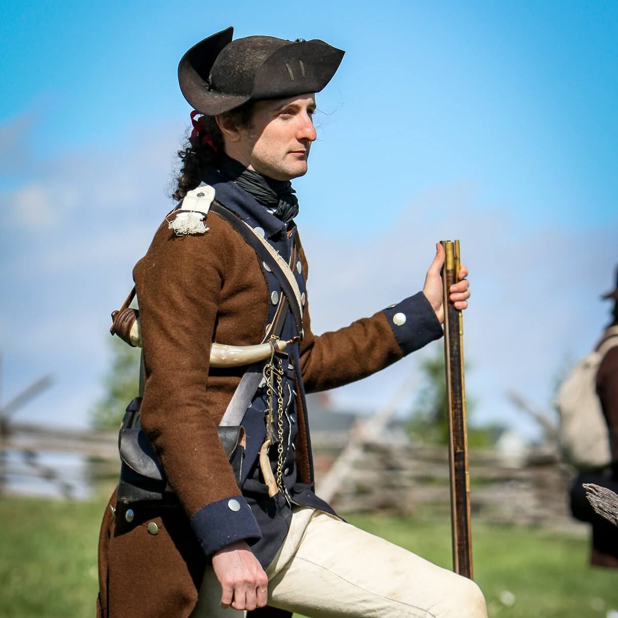 A man dressed in a colonial outfit walks with a stick, showcasing a historical style as he moves forward