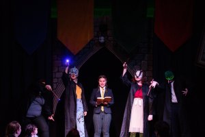 A lively stage scene featuring a person holding a book, surrounded by a group of enthusiastic individuals wearing masks sharing the moment