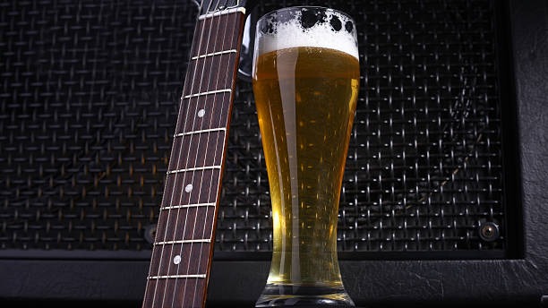 A tall glass of beer next to the neck of an electric guitar in front of a black amplifier
